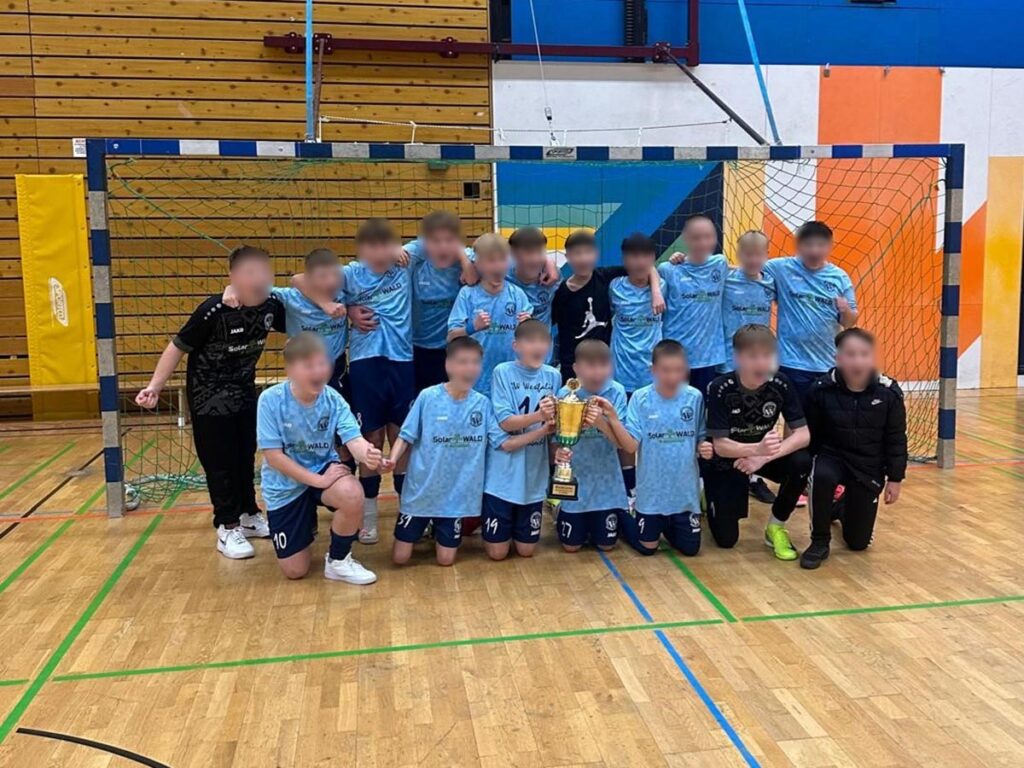 Eine Jugend-Fußballmannschaft mit Pokal, alle lächeln; hellblaue Trikots, Holzwand im Hintergrund.